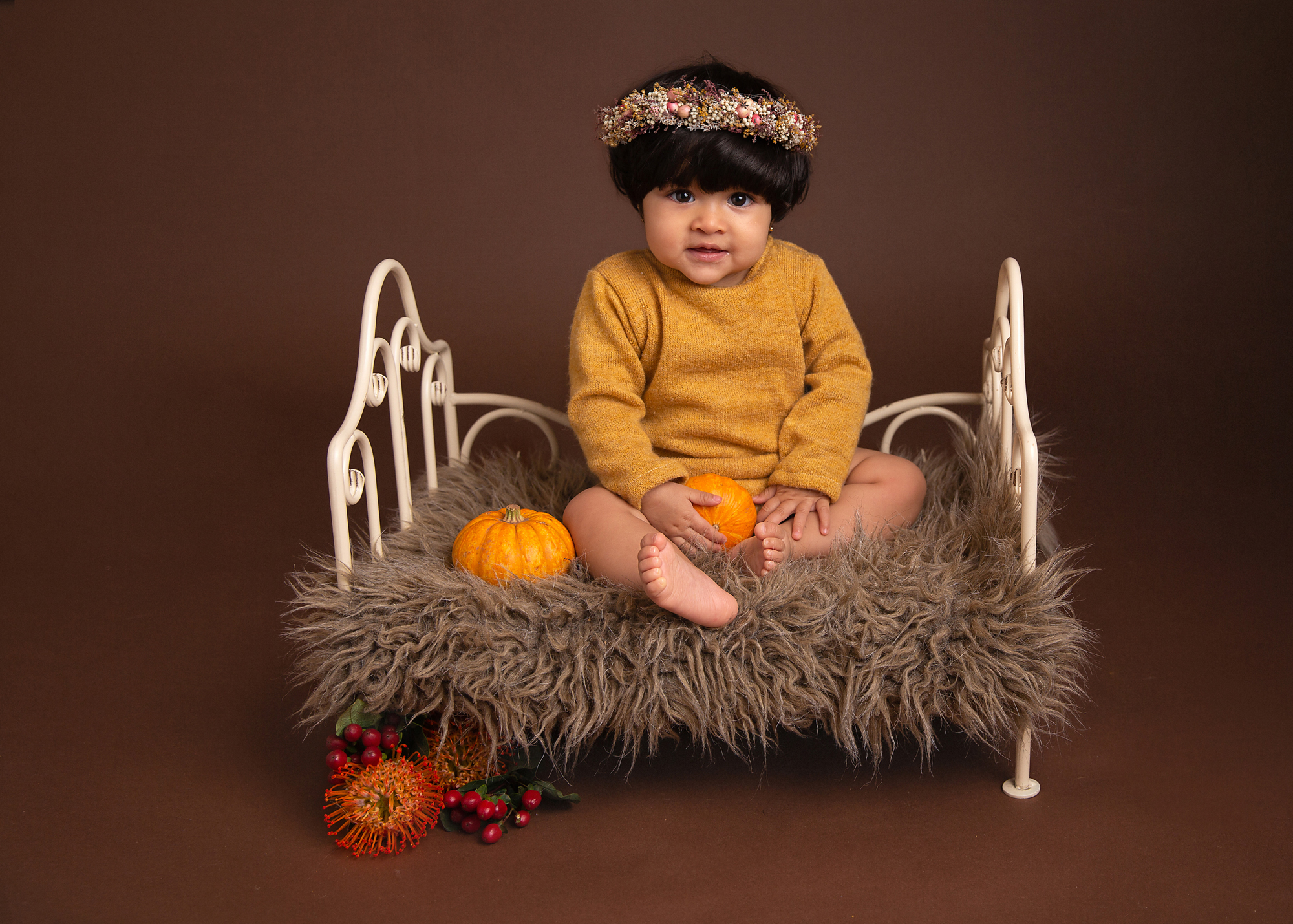 smiling toddler in mustard outfit sitting on a cream metal bed with rustic headband by surrey baby photographer